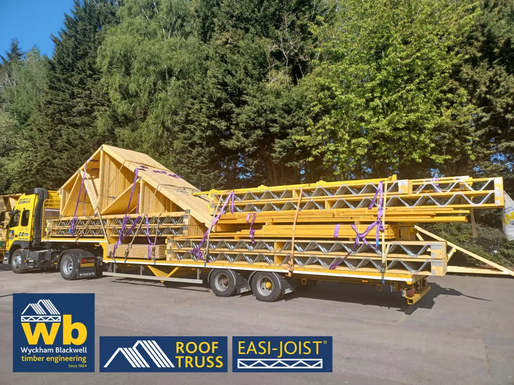 Side angle of timber Roof Truss and Easi-Joist metal web joists loaded and strapped onto a yellow Wyckham Blackwell lorry parked in front of big green trees.