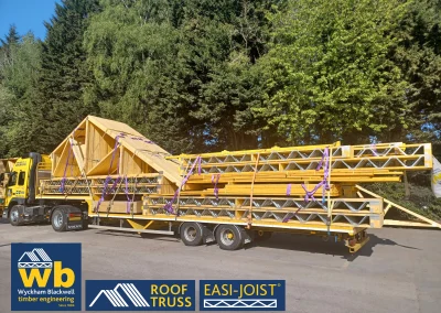 Side angle of timber Roof Truss and Easi-Joist metal web joists loaded and strapped onto a yellow Wyckham Blackwell lorry parked in front of big green trees.