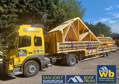 Front side view of timber Roof Truss and Easi-Joist metal web joists loaded and strapped onto a yellow Wyckham Blackwell lorry parked in front of big green trees.