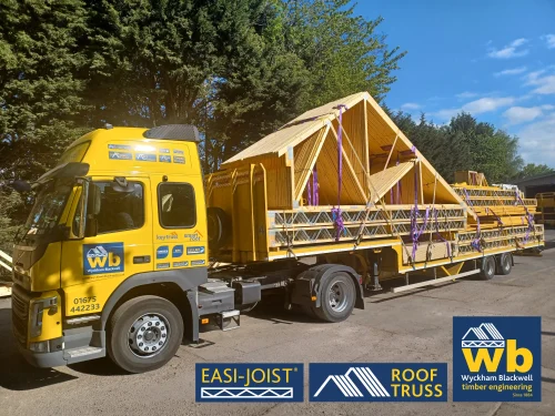 Front side view of timber Roof Truss and Easi-Joist metal web joists loaded and strapped onto a yellow Wyckham Blackwell lorry parked in front of big green trees.