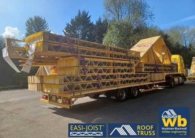 Back angled view of timber Roof Truss and Easi-Joist metal web joists loaded and strapped onto a yellow Wyckham Blackwell lorry parked in front of big green trees.