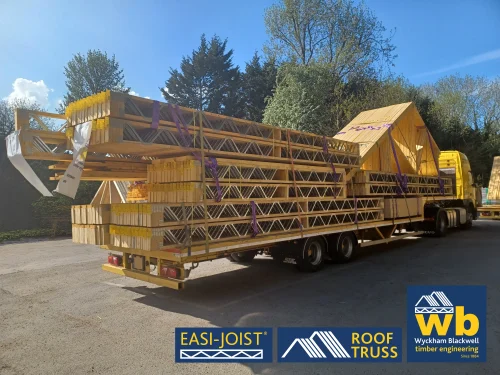 Back angled view of timber Roof Truss and Easi-Joist metal web joists loaded and strapped onto a yellow Wyckham Blackwell lorry parked in front of big green trees.