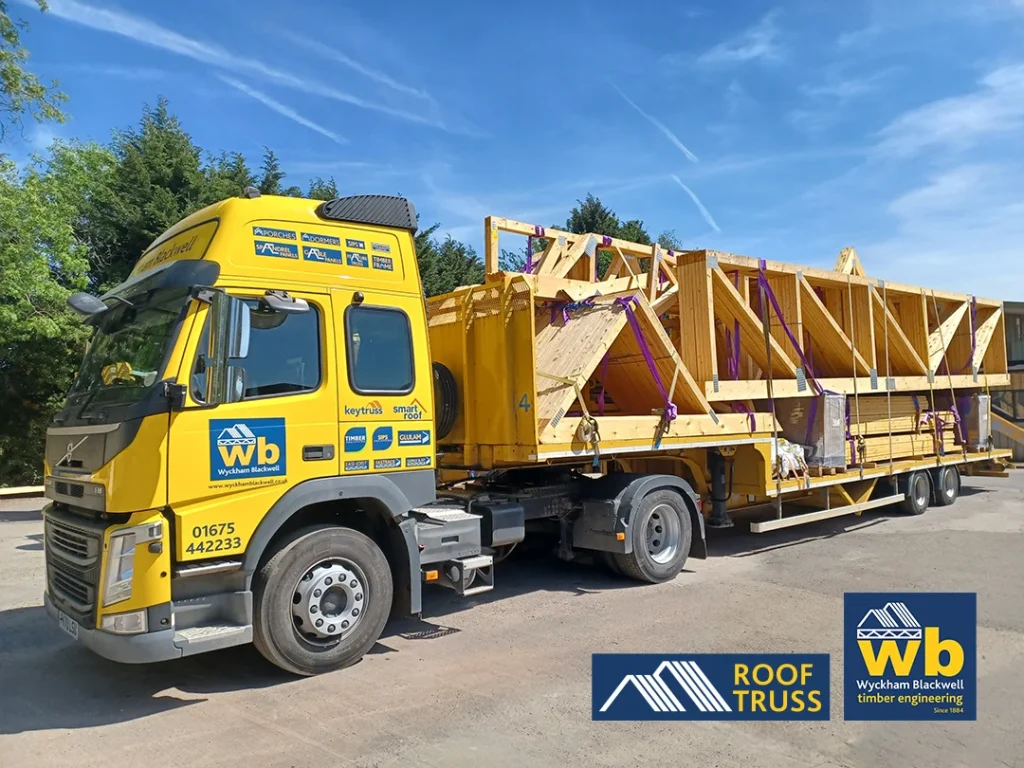 Yellow Wyckham Blackwell flatbed lorry hauling stacked wooden roof trusses secured with purple straps under blue sky.