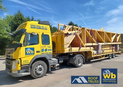 Yellow Wyckham Blackwell flatbed lorry hauling stacked wooden roof trusses secured with purple straps under blue sky.