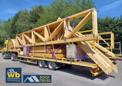 Yellow Wyckham Blackwell flatbed lorry carrying large prefabricated wooden timber roof trusses secured by purple straps, parked outdoors.