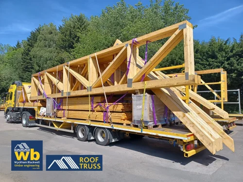 Yellow Wyckham Blackwell flatbed lorry carrying large prefabricated wooden timber roof trusses secured by purple straps, parked outdoors.