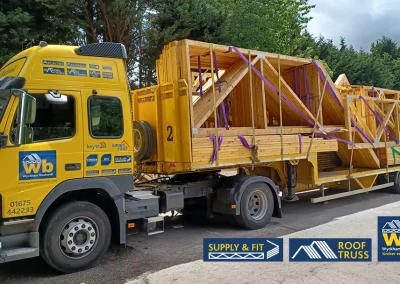 Yellow Wyckham Blackwell lorry hauling stacked wooden roof trusses secured with purple straps on a roadside.