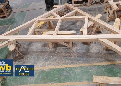 Large oak timber feature truss with intersecting beams resting on sawhorses in a workshop, Wyckham Blackwell logo in bottom right.