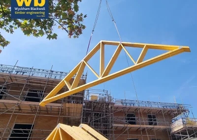 Timber Roof Truss lifted by crane above scaffolding at a construction site, with Wyckham Blackwell logo top left.