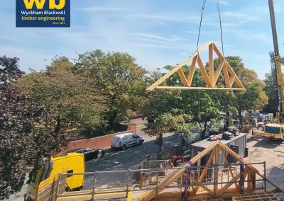 Crane lifts a wooden Roof Truss above a flatbed lorry at a construction site, Wyckham Blackwell logos at top.